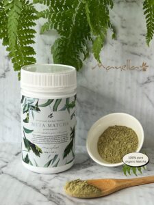 Mayella Meta Matcha container on a white marbled bench with a wooden teaspoon filled with Meta Matcha and a small porcelain bowl on its side with Meta Matcha in. A leafy green fern hangs over behind.