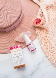 Mayella Olivane Rose Serum lying next to it's pink rose covered box on a rose quartz crystal. Both lying a top a pink sunhat with a slamon mesh shopping tote, pink rose bloom on a white marble stone bench.