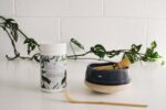 Mayella Meta Matcha on a white bench with a traditional Japanese bamboo scoop spoon and a bamboo whisk sitting inside a blue and white pottery cup.