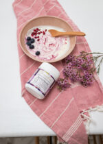 Maella Acai Berry Beautiful Blend lying on its side on a pink cotton tea towel and a sprig of dried flowers next to a stone bowl with coconut yoghurt and Acai Berry Blend stirred in.