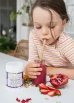 Young girl drinking a Mayella Acai Berry Beautiful Blend Smoothie though a bamboo straw with fresh fruits chopped up on the bench before her.