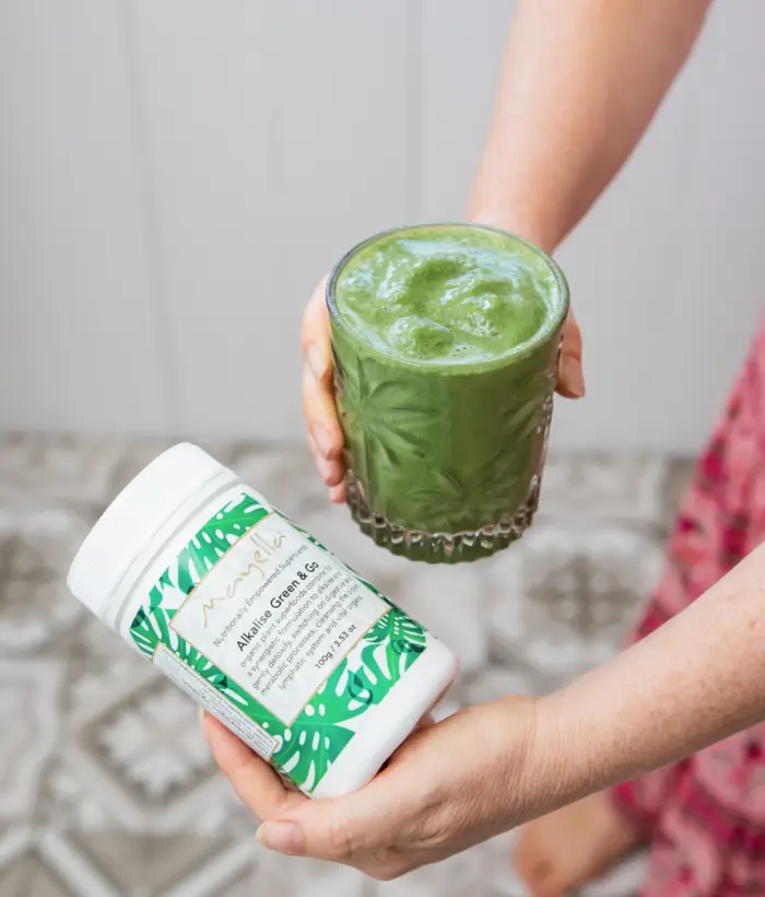 A lady holding Mayella Alkalise Green & Go Blend in her hand next to a thick creamy Alkalise Green & Go Smoothie in a cut crystal glass.