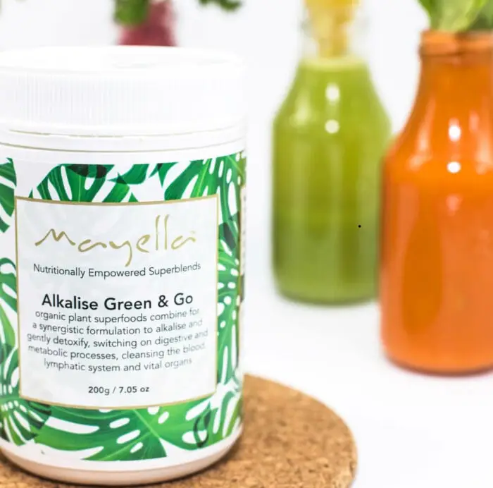 Mayella Alkalise Green & Go Nutritional Blend sitting on a cork mat in front of two galss bottles filled with a carrot juice and a green apple juice.