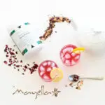 Mayella Beauty & Brains Herbal Tea container l;ying on its side with a white teapot pouring the loose leaf tea out in a dance around the tea container and two glasses filled with the pink tea, ice cubes and a slice of lemon on side of the glass.