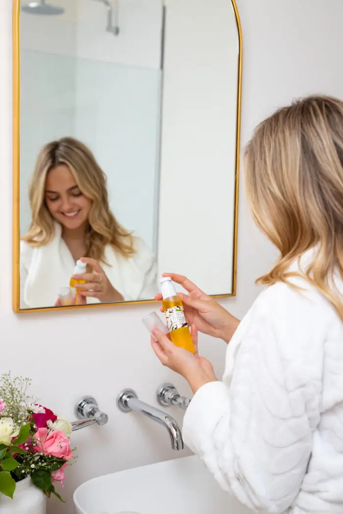 A blonde lady wearing a white towellng bath robe, in her bathroom in front of the mirror with Mayella Cleansing Serum in her hands.