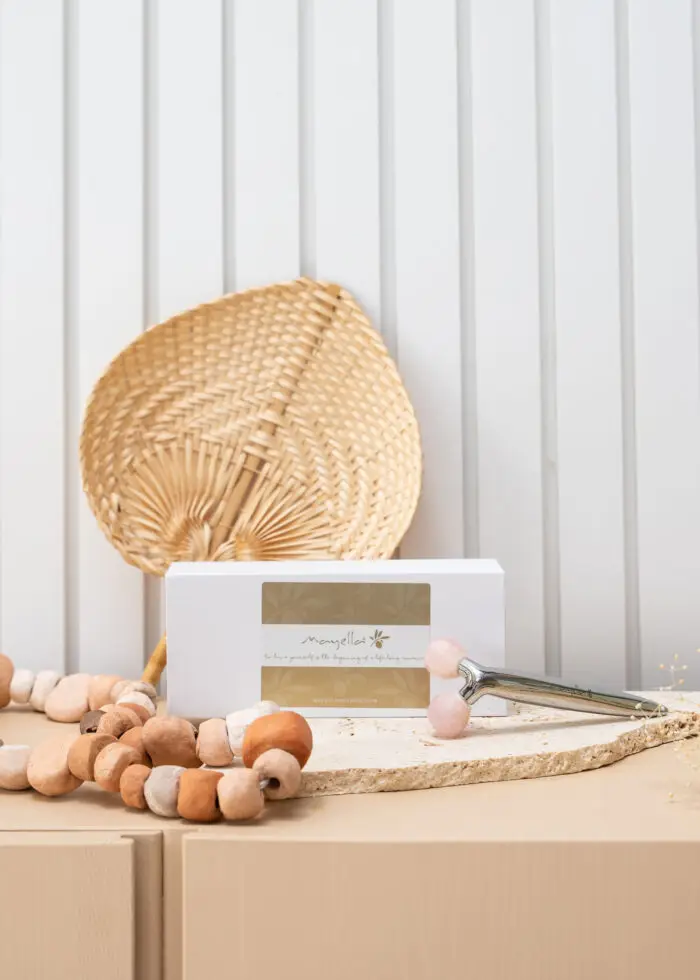 A cane woven fan in the background behind the white luxe box of Mayella Facial defining roller with two faceted rose quartz balls lying on a disc of cream natural stone. A string of natural toned wooden beads lies to the front left.