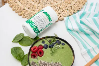 Mayella Alkalise Green & Go lying on its side next to a green smoothie bowl made with Mayella Alkalise Green & Go on a white table with green & white striped tea towel.