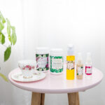 Mayella Beauty & Brains Herbal Tisane, Alkalise Green & Go, Cleansing Serum, Multi Vitamin Whip and Olivane Repair Serum - Infusion Rose are sitting with a white & floral porcelain china tea cup and saucer on a pink side table next to a green indoor palnt.