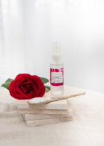 Mayella Olivane Rose sitting on a stack of natural stone tiles with a handcrafted pottery spoon lying across in front. A deep red rose lies to the left of the bottle.