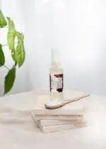 Mayella Olivane Spice sitting on a stack of natural stone tiles with a handcrafted pottery spoon lying across in front. A leafy green houseplant lies to the left of the bottle.