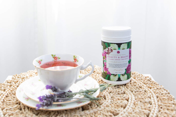 A container of Mayella Beauty & Brains Herbal tea sitting on a delicately woven rope mat next to a china cup and saucer with filled with the rosy pink Beauty & brains tea.