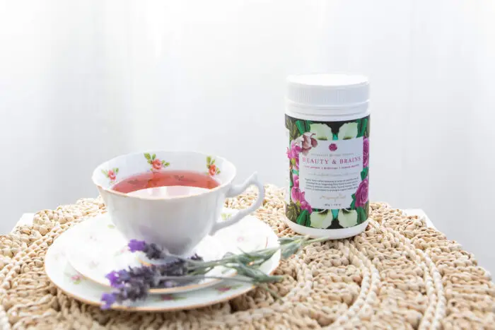 A container of Mayella Beauty & Brains Herbal tea sitting on a delicately woven rope mat next to a china cup and saucer with filled with the rosy pink Beauty & brains tea.