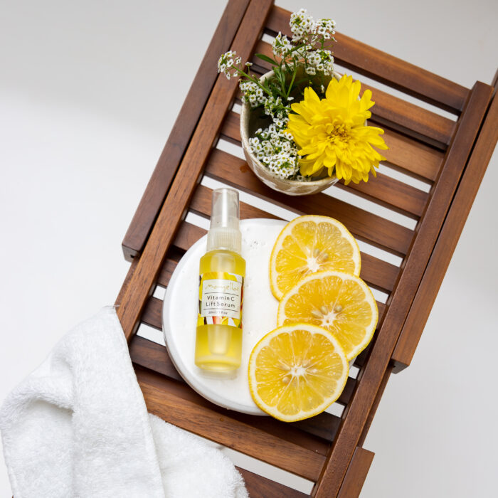 Mayella Vitamin C Lift Serum 30ml lying on a white plate with 3 round slices of Lemon. The plate is on a wooden rack with a bunch of yellow and white assorted flowers top and a white fluffy towel in the bottom left slung across the rack.