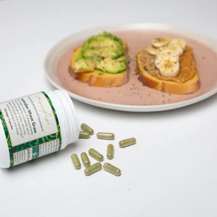 Mayella wheatgrass capsules container lies on its side with the green capsules spilling out across the table in front of a pink plate with two pieces of toast on it. One piece has avocado and one piece has peanut butter and banana sliced on top.