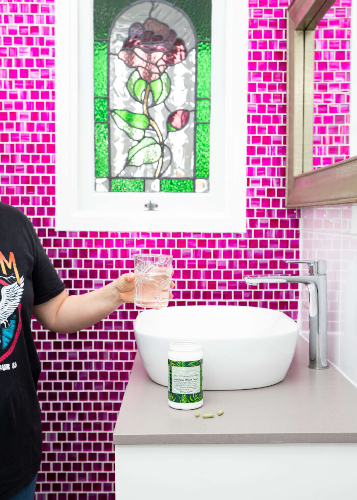 A lady stands with her arm outstretched holding a glass of water in her left hand over a white bathroom basin. a container of Mayella Wheatgrass 150 capsules stands on the bathroom bench with a handful of capsules beside the container. A bright pink tiled wall and green glass leadlight window are in the background.