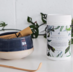 Mayella Meta Matcha on a white bench with a traditional Japanese bamboo scoop spoon and a bamboo whisk sitting inside a blue and white pottery cup.