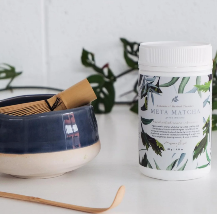 Mayella Meta Matcha on a white bench with a traditional Japanese bamboo scoop spoon and a bamboo whisk sitting inside a blue and white pottery cup.