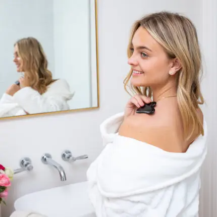 A lady standing dressed in a white fluffy towelling robe in front of a mirror massaging her shoulder with a Mayella Black Obsidian Gua Sha