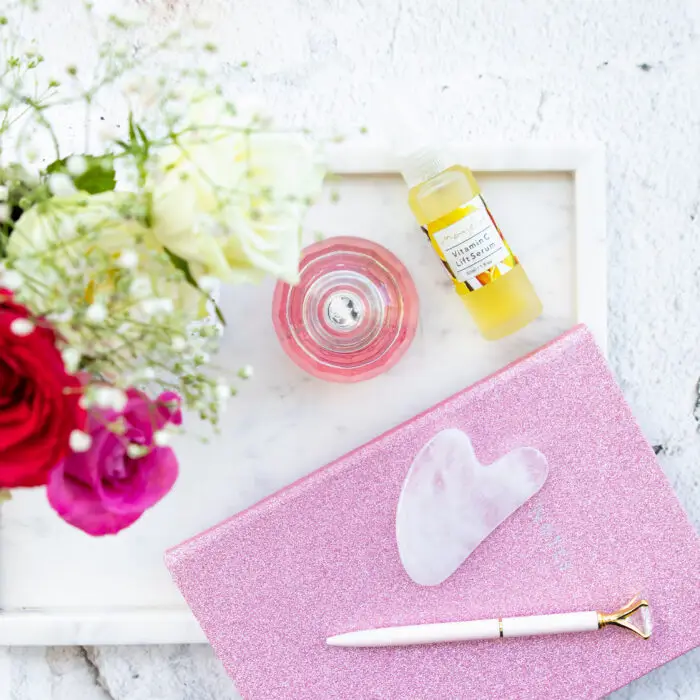 A flatlay view of Mayella Clear Quartz Gua Sha lying on a pink journal with a white handled writin pen. Mayella Vitamin C Lift Serrum is standing next to the journal with a vase of roses and a crystal perfume bottle