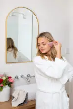 A lady standing dressed in a white fluffy towelling robe in front of a mirror massaging her shoulder with a Mayella Rose Quartz Gua Sha