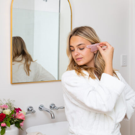 A lady standing dressed in a white fluffy towelling robe in front of a mirror massaging her shoulder with a Mayella Rose Quartz Gua Sha