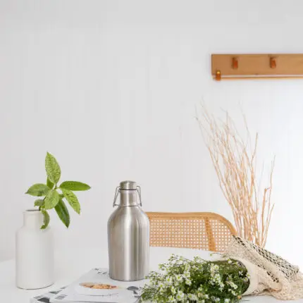 Mayella 1.9L stainless steel flask sitting on a white table with cane chair in background and a calming green house plants to left, fresh bought white flowers lying on table to the right.