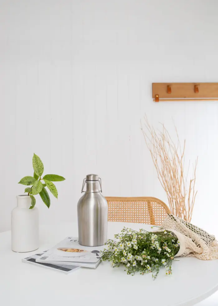 Mayella 1.9L stainless steel flask sitting on a white table with cane chair in background and a calming green house plants to left, fresh bought white flowers lying on table to the right.