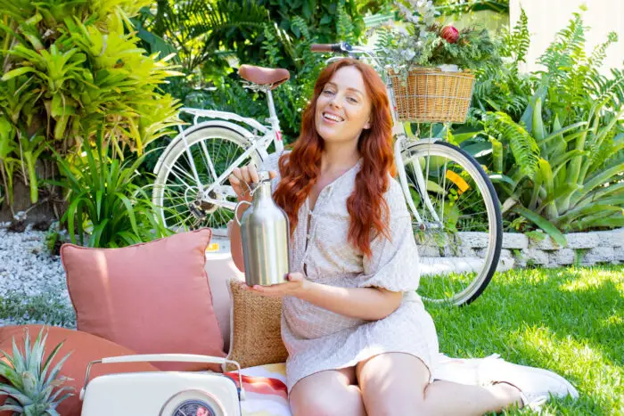 A lush garden setting with a white cruiser bicycle on the grass and a lady with long red hair kneeling on a picnic blanket. A big pink cushion, old style transistor radio and assorted fresh fruits for her picnic as she holds Mayella Stainless Steel flask in her hands.