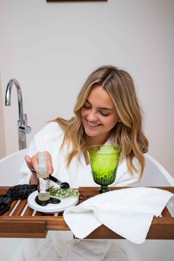 A lady in a white fluffy bathrobe sitting in the bath picking up a Mayella Black Obsidian Roller with MOJO Spice on the wooden bath rack next to a green cut glass drink