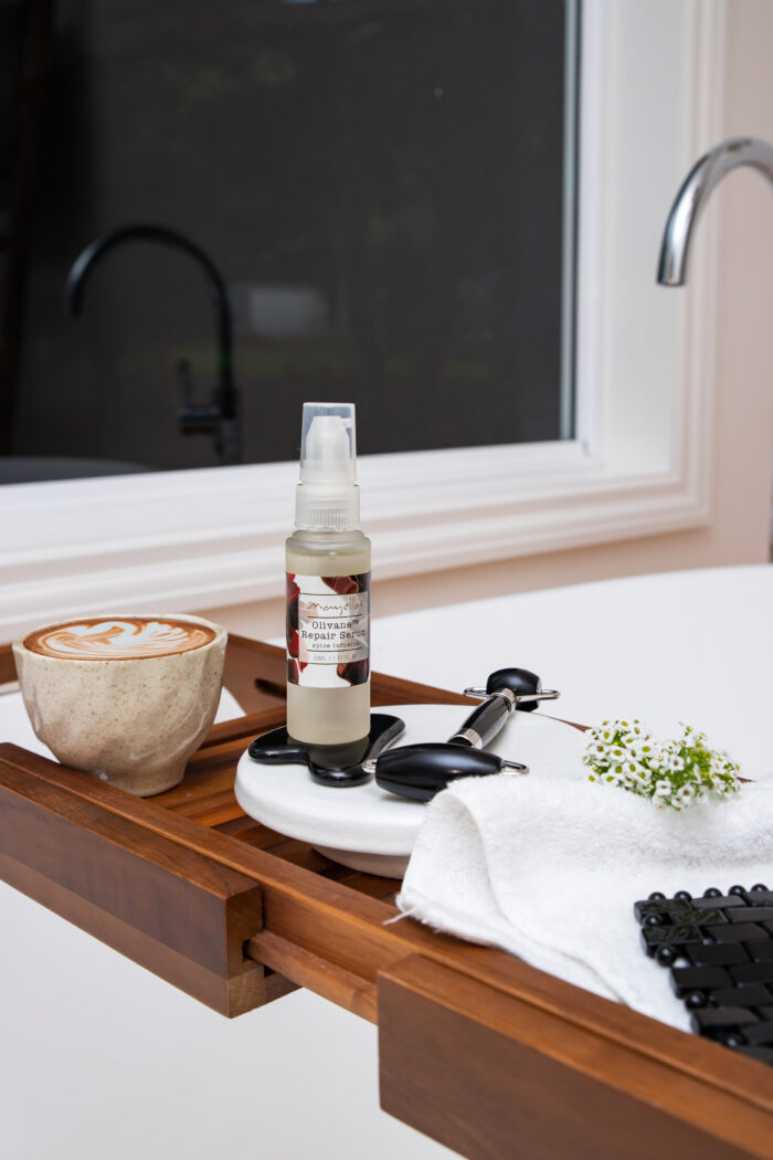 An evening bath & Skin care ritual with a cappacino in a clay cup sitting next to Mayella MOJO Spice Serum Mayella Black Obsidian Gua Sha & Roller on a wooden rack with a white fluffy towel thrown next to these skin care products